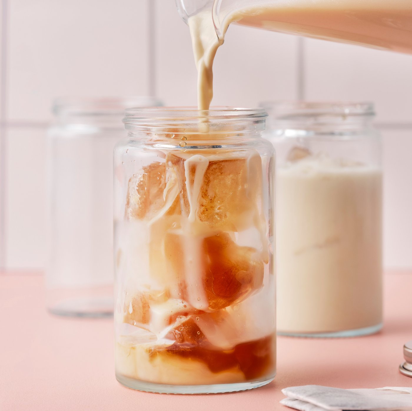 A hand is pouring oat drink for coffee from a jug into a glass jar filled with ice cubes made of water and FLÖJT black tea.