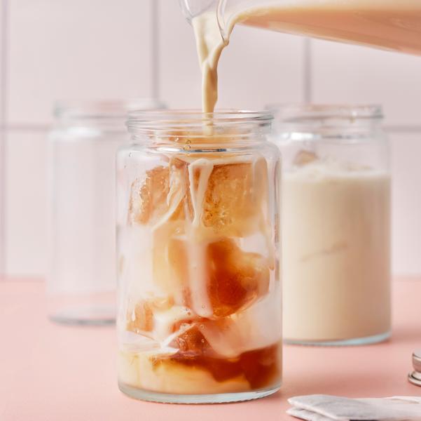 A hand is pouring oat drink for coffee from a jug into a glass jar filled with ice cubes made of water and FLÖJT black tea.