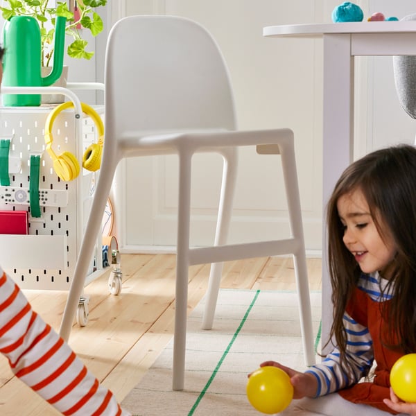 A white URBAN junior chair stands next to children playing with colorful balls on a wooden floor in a bright room.