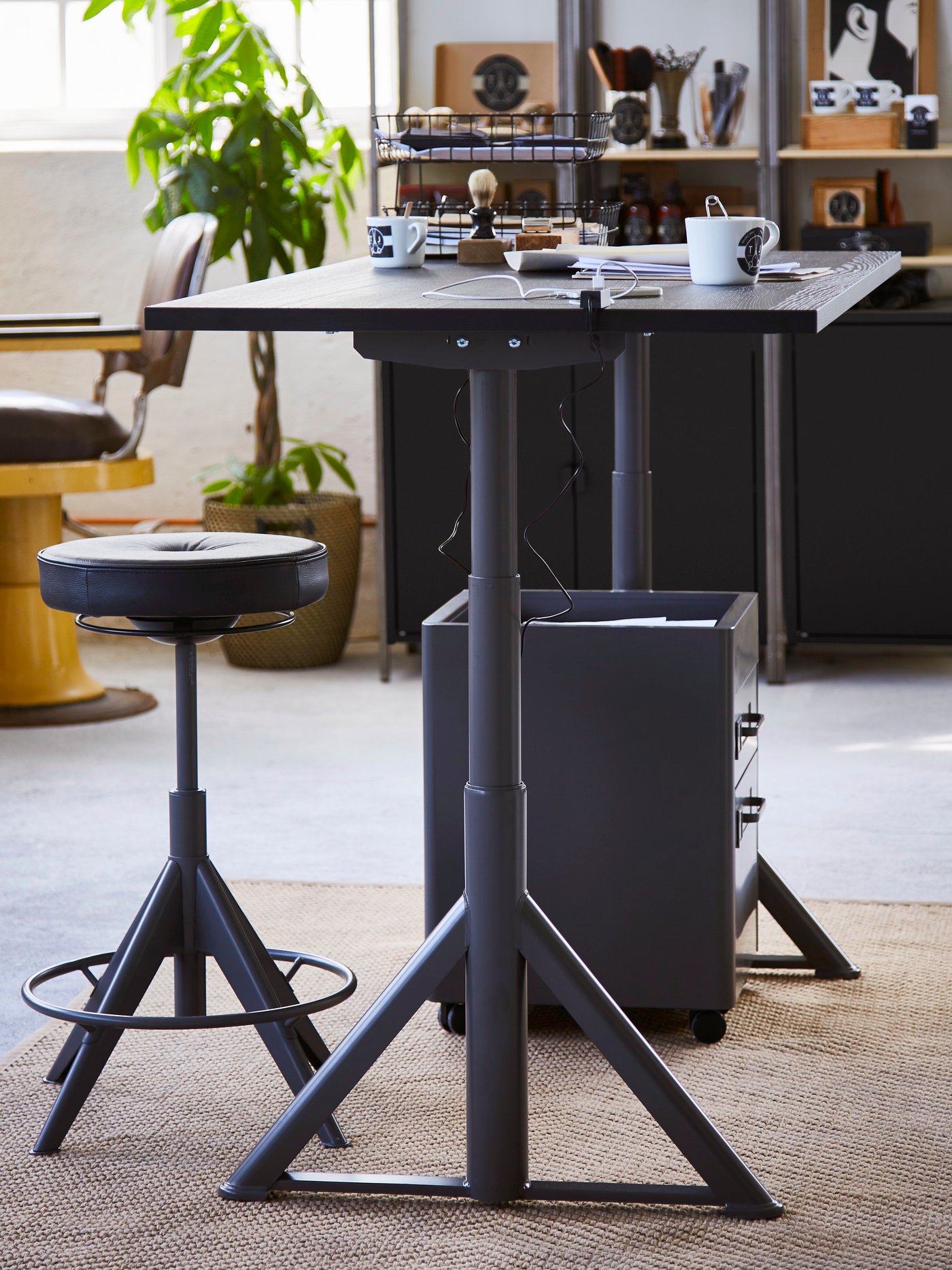Detalle de un espacio de trabajo en el que se encuentra un escritorio negro rectangular de altura regulable y diseño minimalista con un soporte activo para trabajar de pie/sentado.