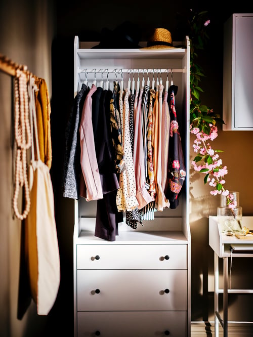 Weißer offener HAUGA Kleiderschrank mit 3 Schubladen, Kleidern an einer Kleiderstange und einem Schreibtisch rechts daneben, auf dem eine Vase mit Blumen steht.