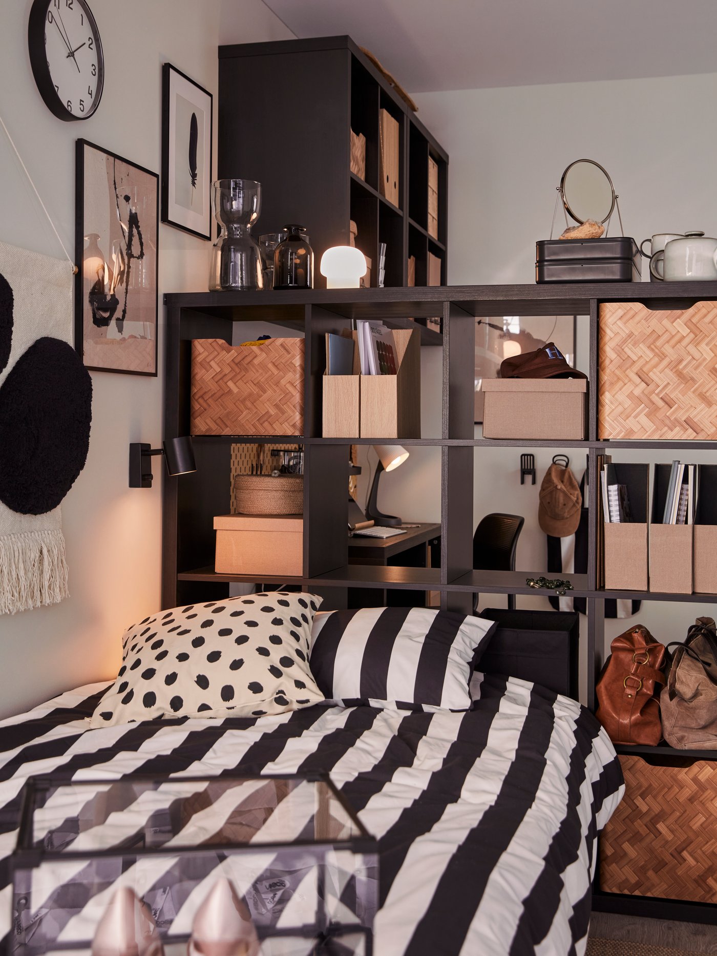 Dans une chambre d’étudiant, une grande étagère KALLAX noir-brun agrémentée d’accessoires et de boîtes fait office de tête de lit.