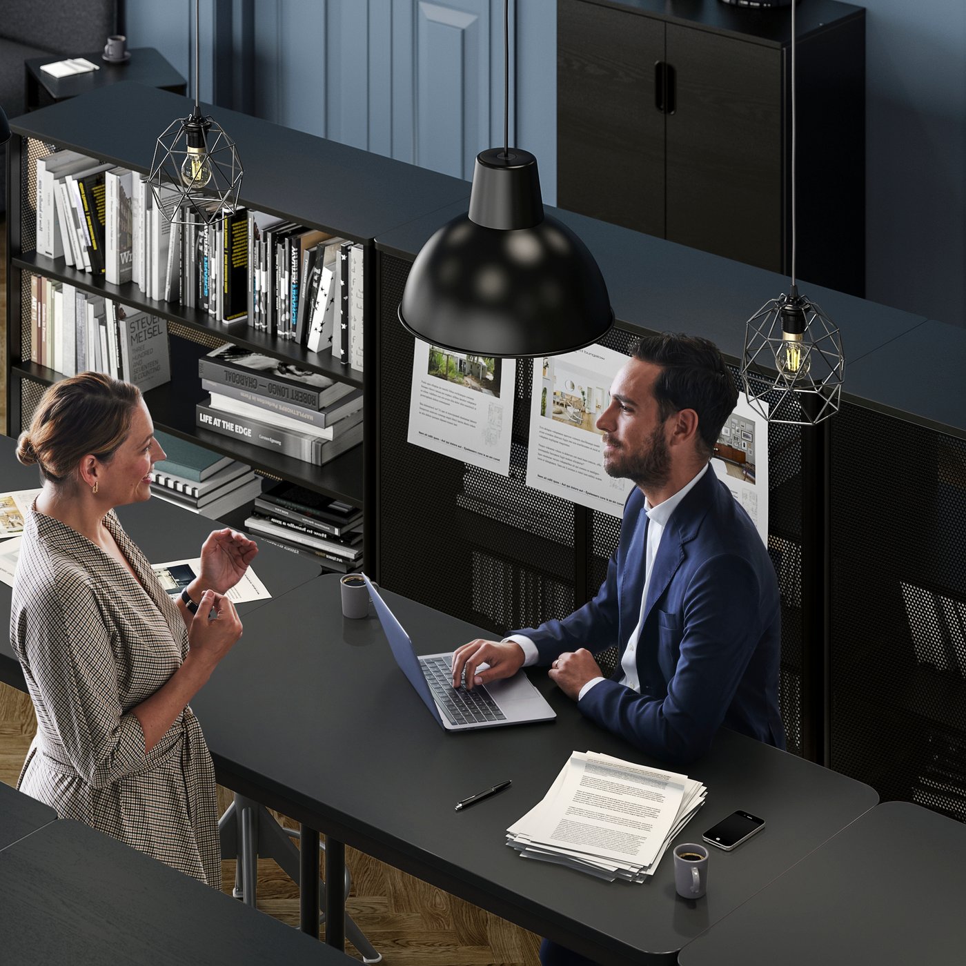 Un uomo in giacca e cravatta in piedi vicino a una scrivania in ufficio, intento a scrivere su un laptop mentre conversa con una donna seduta dall'altro lato della scrivania.