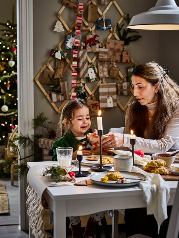 Mutter und kleine Tochter frühstücken an einem weißen Tisch mit FULLTALIG-Kerzenhalter und GLADELIG-Tasse in weihnachtlicher Umgebung.