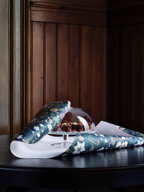 An ARV BRÖLLOP serving stand with lid partially covered by a gift wrap roll, displayed on a round table.