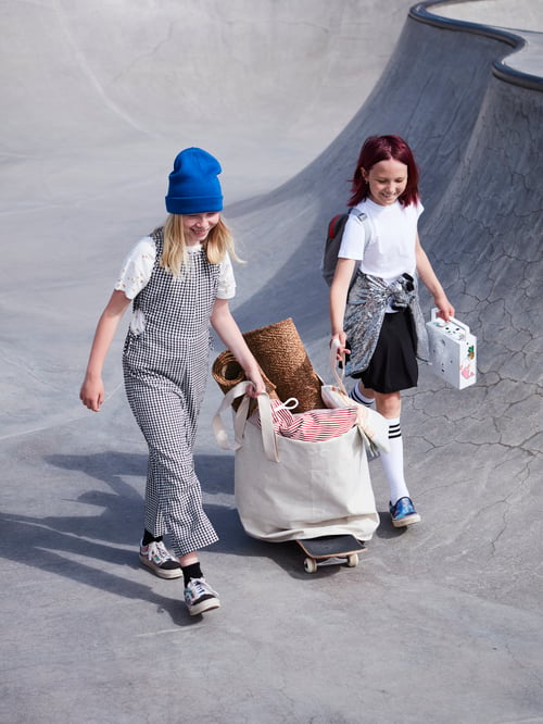 Două fete de aproximativ zece ani care transportă un coș de rufe în care se află un covor LYDERSHOLM și perne, cu ajutorul unui skateboard, într-un parc.