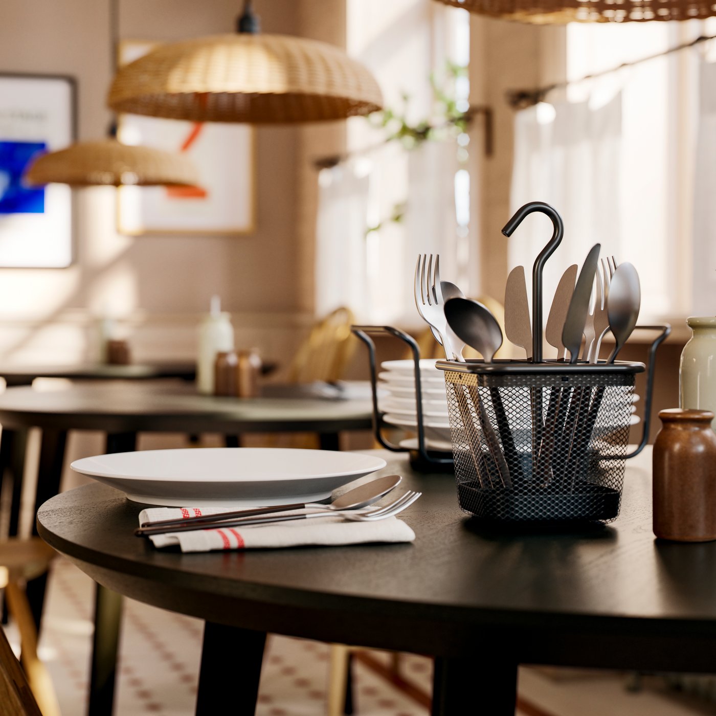 Sul tavolo un portaposate UPPDATERA in maglia metallica color antracite, accanto a un servizio da tavola apparecchiato e una pila di piatti.