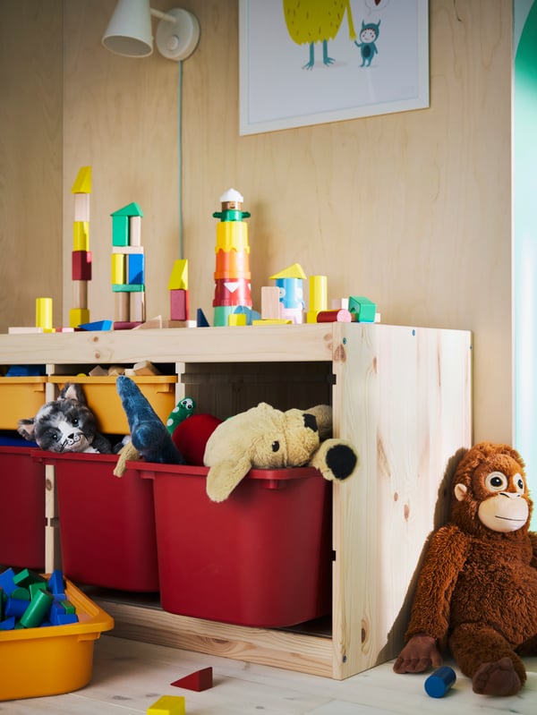 Meuble de rangement TROFAST avec de nombreux jouets sur le dessus et dans des boîtes en plastique rouge/orange. Un singe en peluche appuyé contre lui.