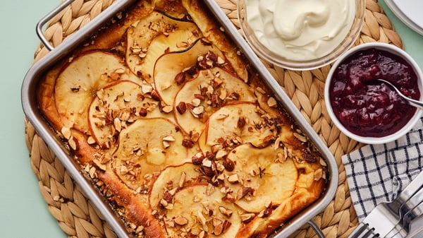 A golden-brown oven pancake with apples and almonds has been baked in a metal baking tray that is placed on a green surface.