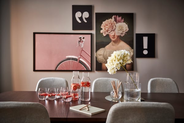 Konferensrum med ett mörkt träbord och grå stolar. På bordet står två karaffer med vatten, glas och en vas med vita blommor. På väggen bakom syns tavlor med citattecken, ett porträtt av en kvinna med blommor över ansiktet, ett utropstecken och ett rosa cykelhjul.