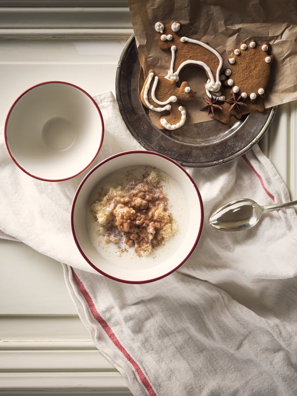 Due ciotole bianche con bordo rosso della collezione VINTER 2020, di cui una piena di porridge, su un tavolo, accanto a un piatto di biscotti allo zenzero.