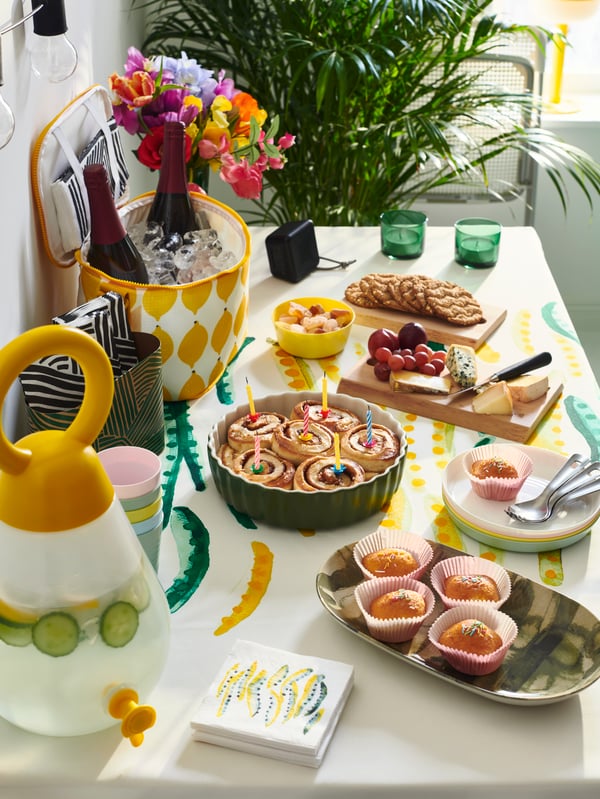 A buffet is set for a party. Platters of cinnamon buns, muffins, cheese and crackers are laid out on top of a tablecloth.