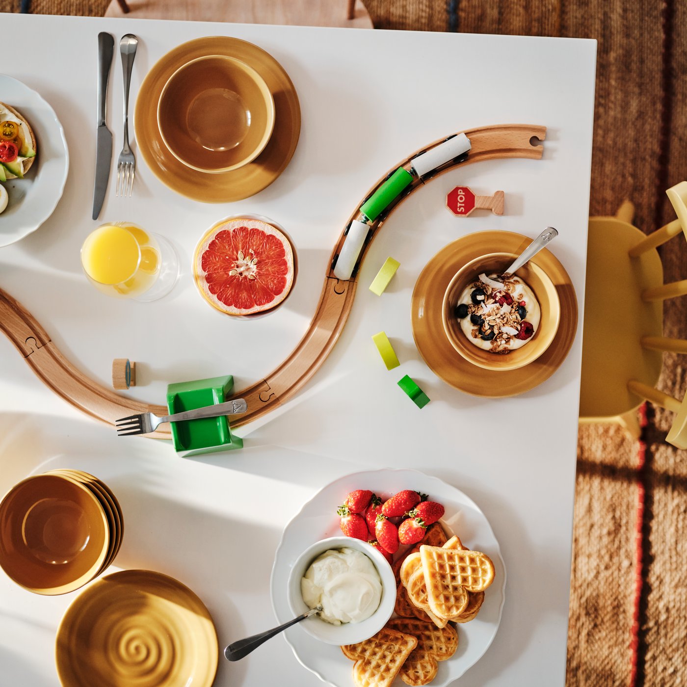 Top shot of a breakfast table set with a plate of waffles and several bowls. A toy train track snakes between them.