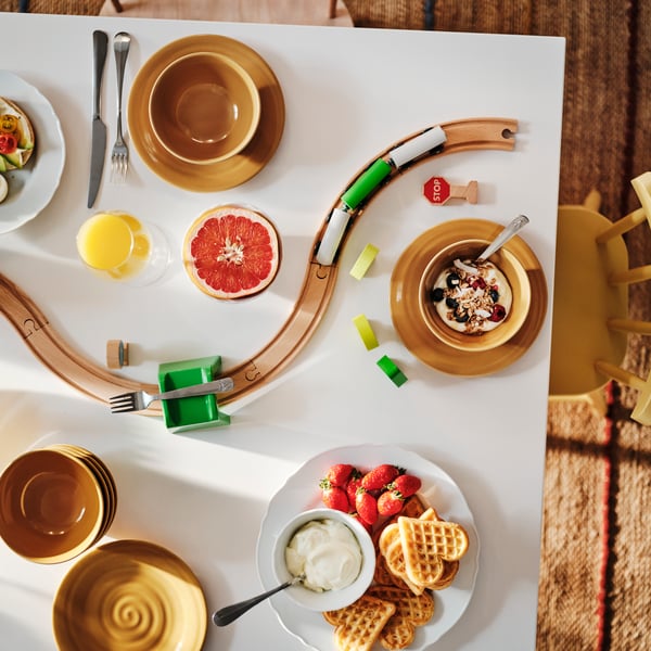 Bovenaanzicht van een ontbijttafel met een bord wafels en verschillende kommen. Een speelgoedtreinspoor slingert zich ertussen.