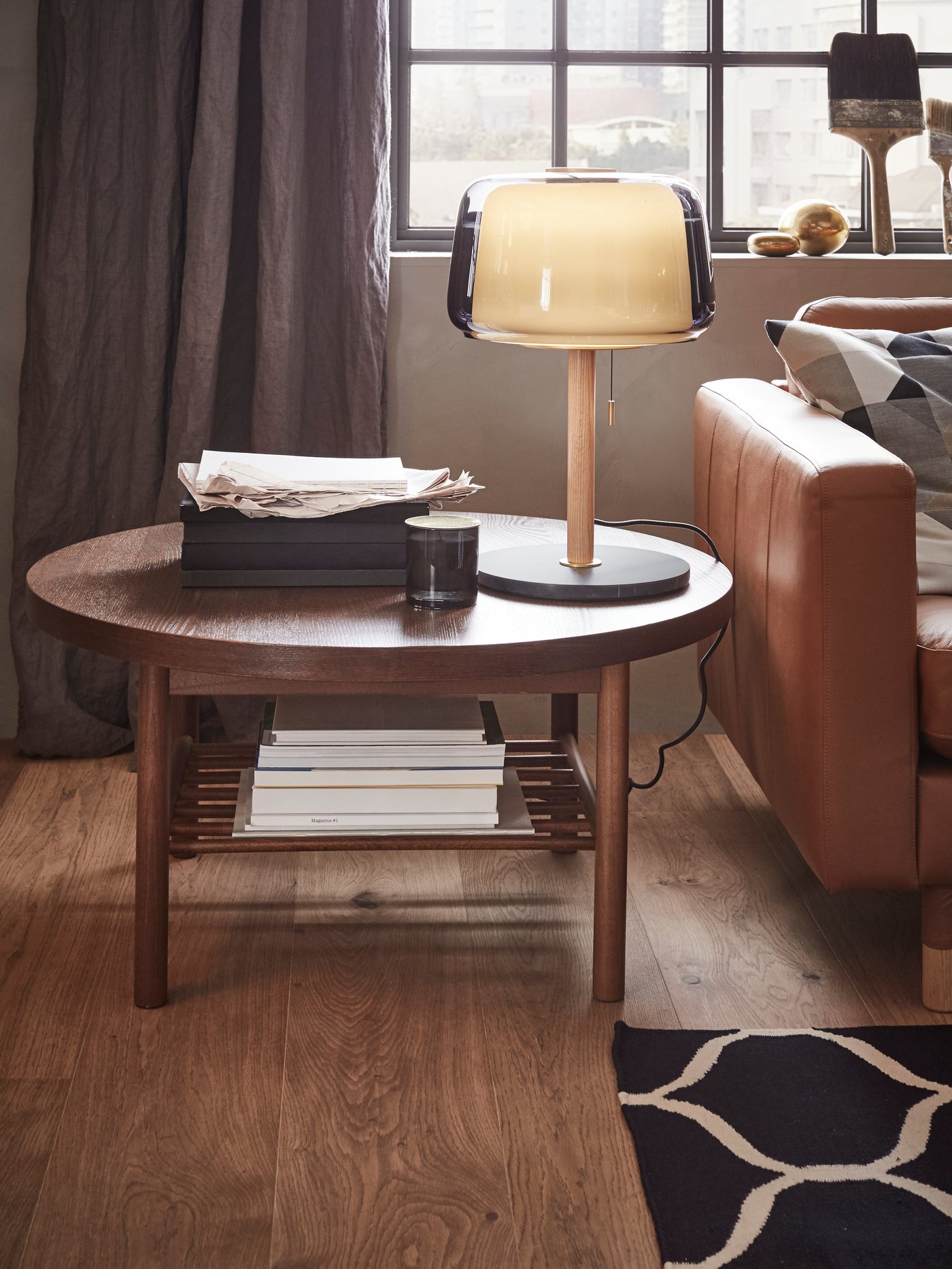 Et rundt sofabord i mørkt træ med en glaslampe står ved siden af en cognac-farvet lædersofa.