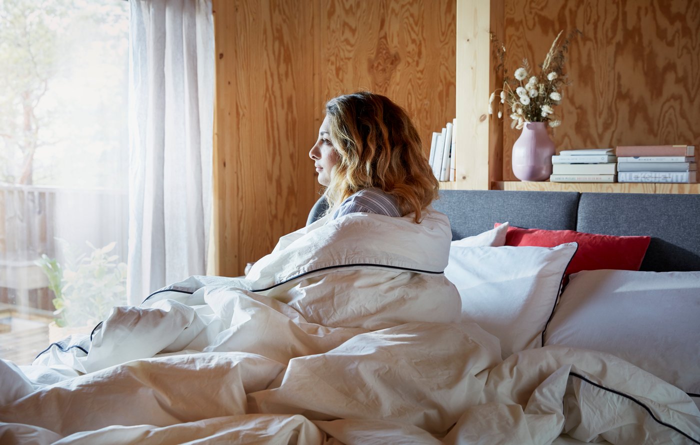 Una mujer joven sentada sobre una cama IKEA mirando hacia una ventana. La mujer está cubierta con un edredón.