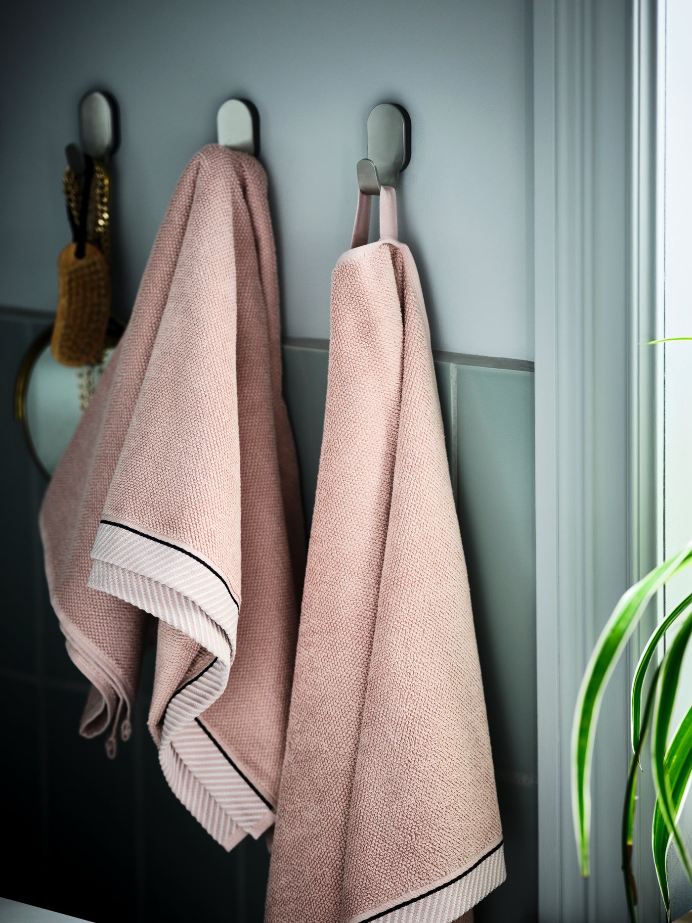 Three hooks on the wall of a green-tiled bathroom hold light-pink VIKFJÄRD hand towels and other bathroom accessories.