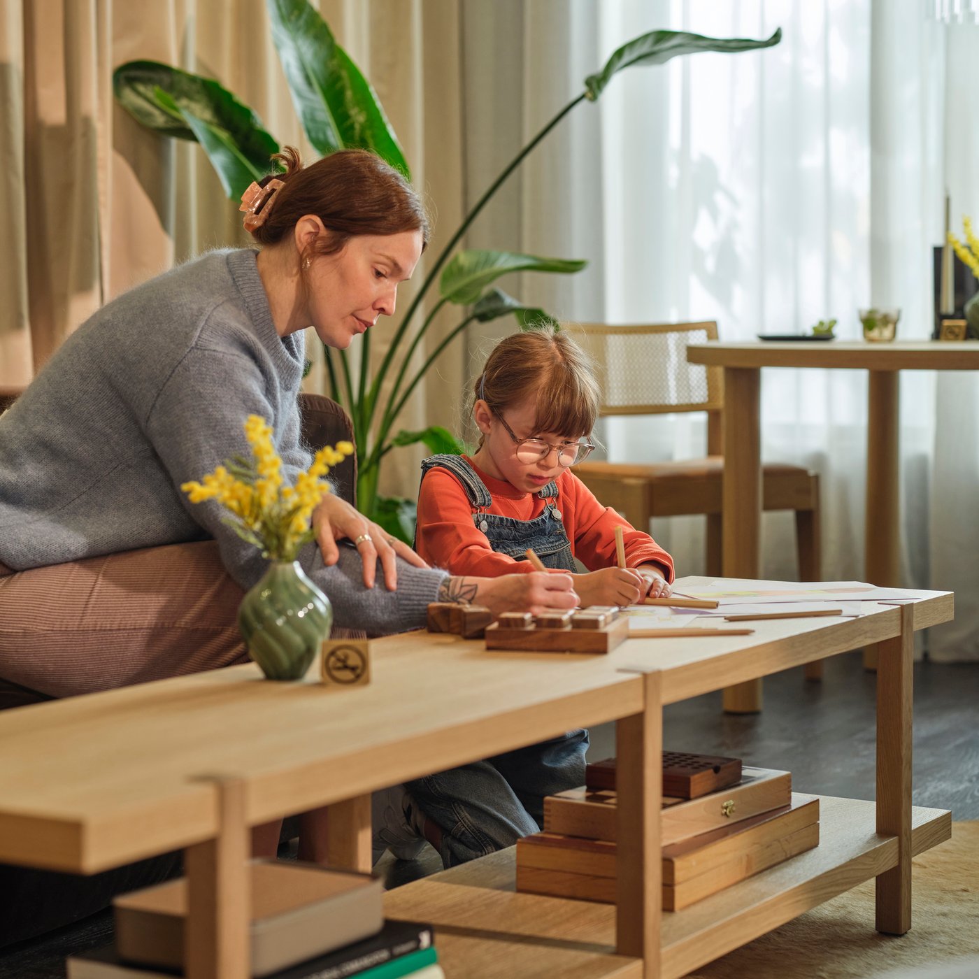 Adult guiding child drawing at a low table in a bright, cozy room with plants and wood furniture.