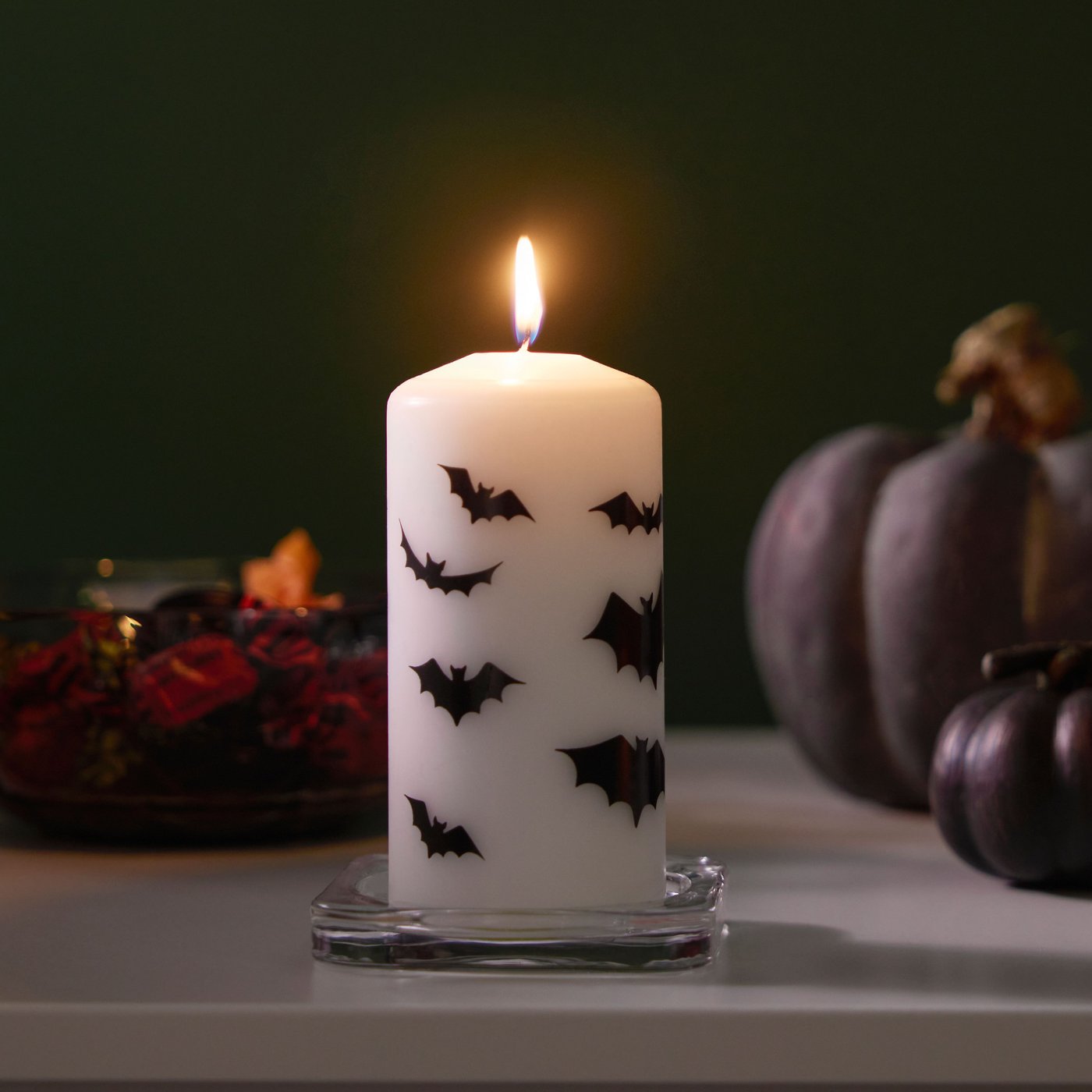 Egy fehér/denevér HÖSTAGILLE illatmentes gyertya ég lánggal az előtérben. Fekete díszek láthatók a háttérben.