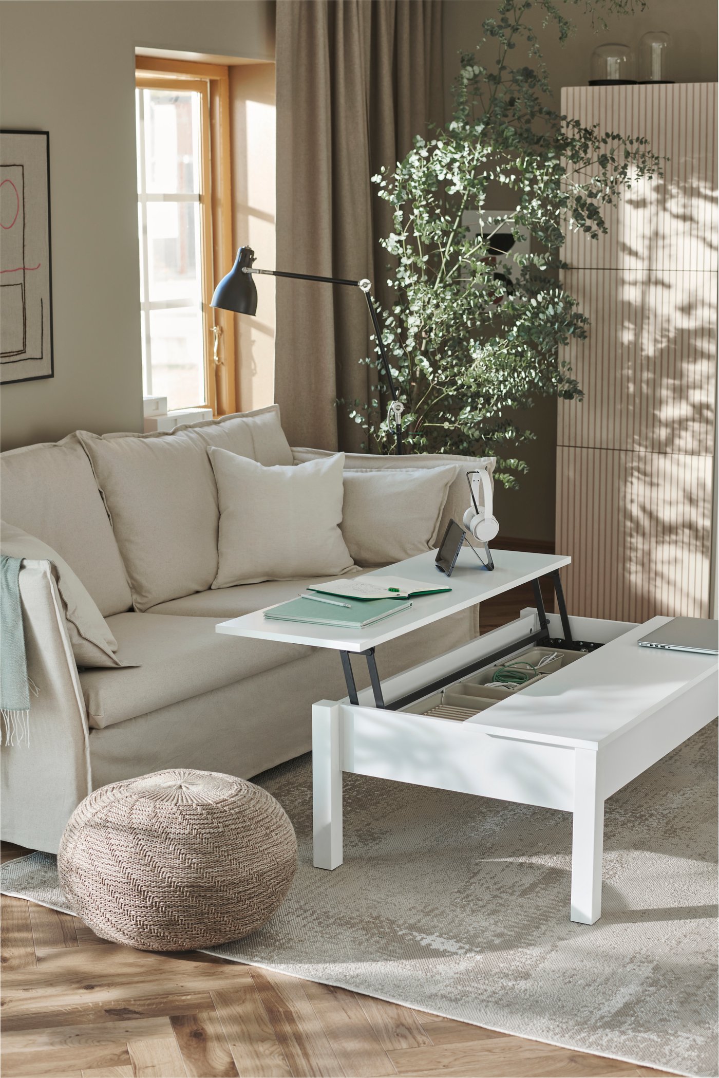 Una mesa de centro TRULSTORP blanca con espacio de almacenaje oculto, con una superficie desplegada. Sobre la superficie elevada hay cuadernos y un soporte para aparatos electrónicos. La mesa está en medio de un living en tonos beige, con una alfombra RODELUND, un sillón BACKSÄLEN y un puf SANDARED. 