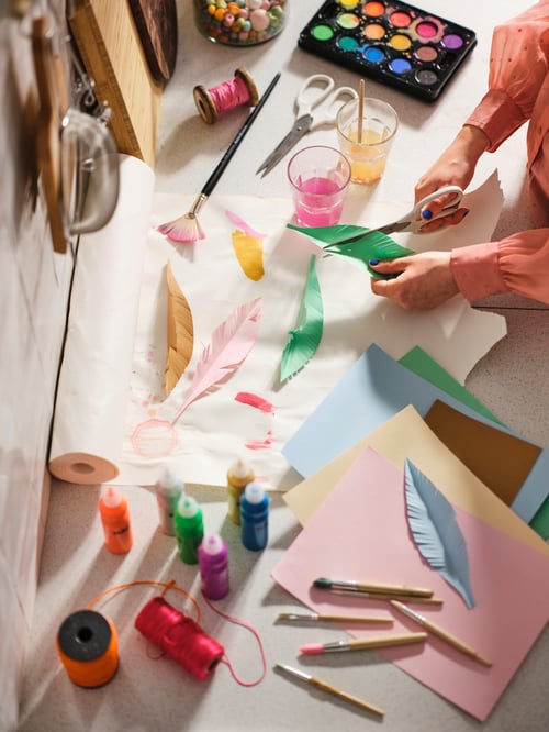 Baby and children room with a child cutting colourful paper into a feather shape, a watercolour box and a drawing paper roll.