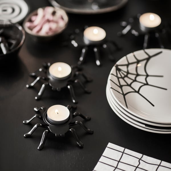 Some KUSTFYR tealight holders shaped like spiders are placed on a black table next to some KUSTFYR side plates.
