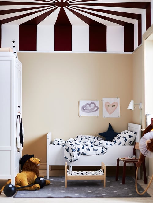 A child’s room with a white bed covered in URSKOG bedlinen. There’s a light on the wall and a DJUNGELSKOG soft lion on the floor.