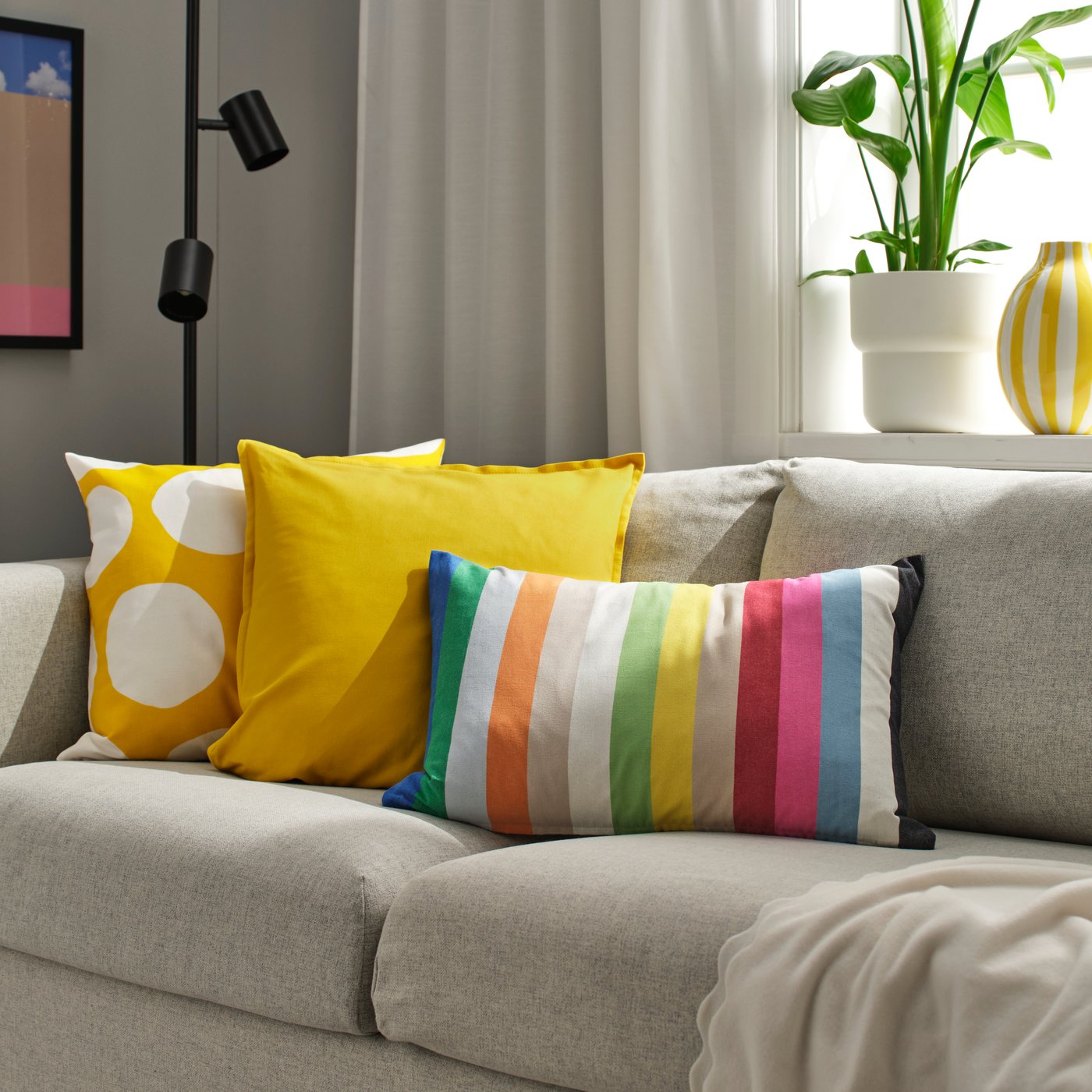 Auf einem beigen Sofa befinden sich ein gelbes BLÅMUNKAR Kissen mit weißen Punkten, ein gelbes GURLI Kissen aus Baumwolle und ein längliches, bunt gestreiftes BÄCKMÄRKEN Kissen.