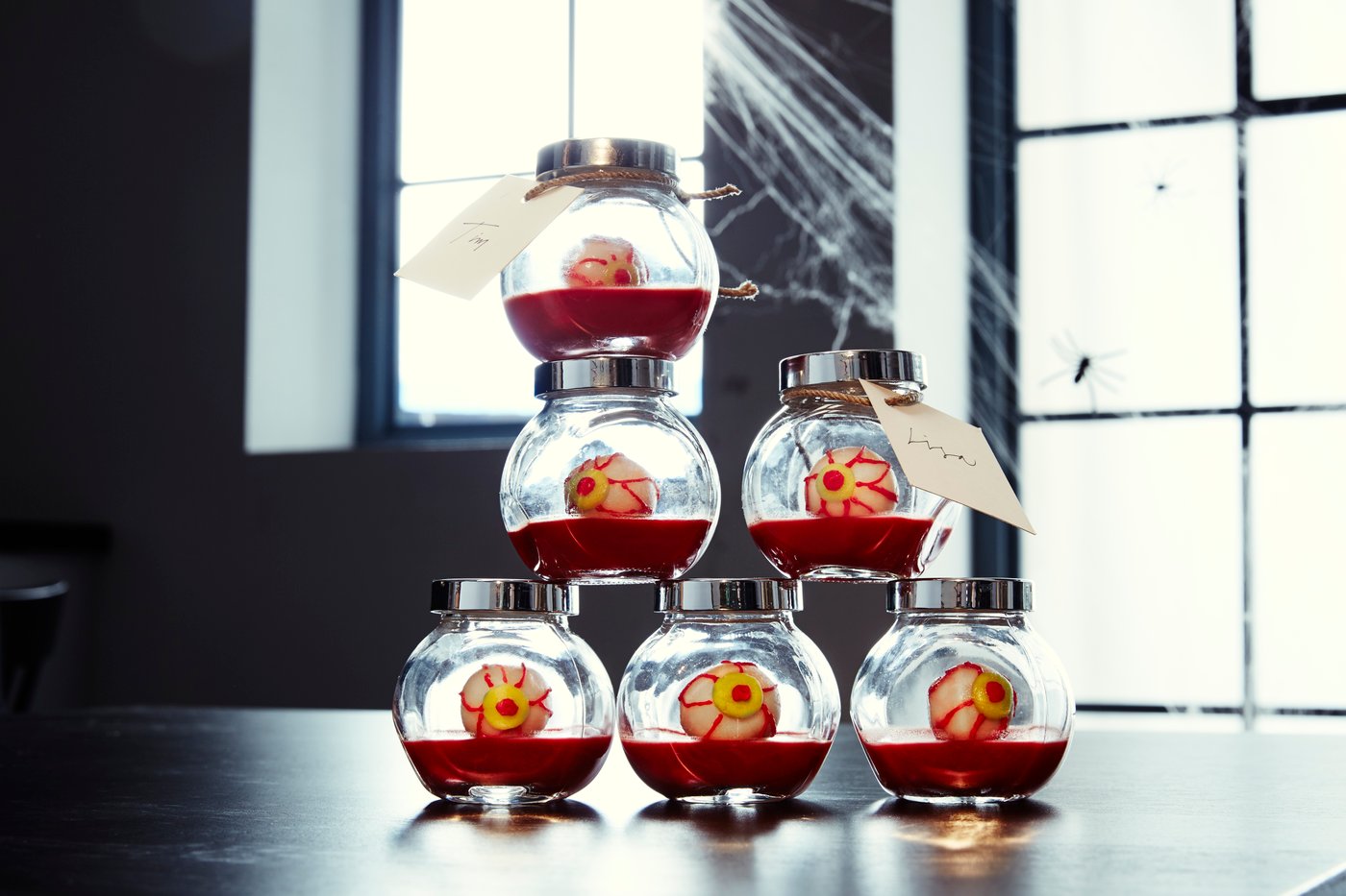 Seks krydderglass fylt med falsk blod og øyeepler sitter på et bord stablet med spindelvev rundt dem