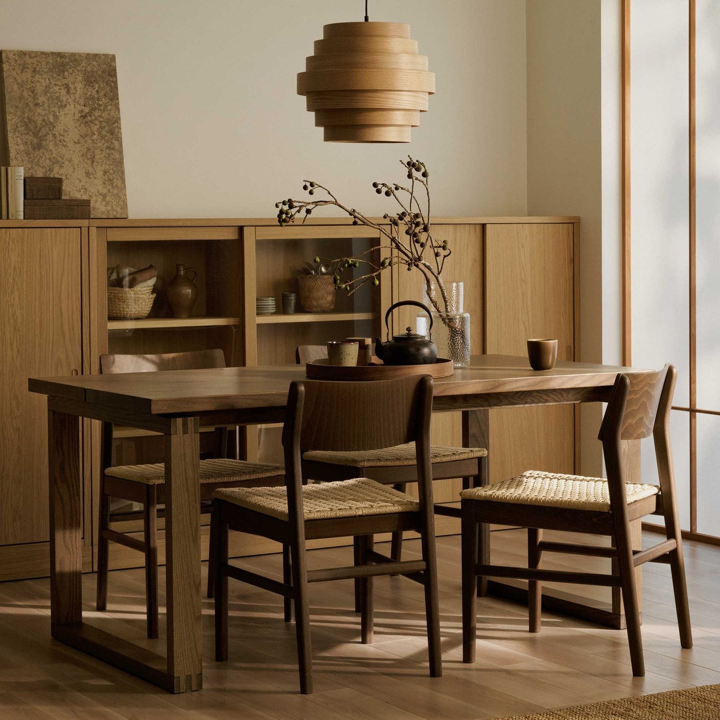 A dining room featuring a long, rectangular wooden dining table with four matching wooden chairs that have woven seats. Above the table hangs a unique, layered wooden pendant light. In the background, there's a light-coloured wooden cabinet with open shelves displaying various decorative items and closed sections. To the right, a tall screen with translucent panels allows soft light to filter in. A branch with dark berries in a vase sits on the table, along with a dark teapot and small cups on a wooden tray, and a single brown mug. The room has a warm, natural aesthetic with light-coloured walls and wooden flooring.