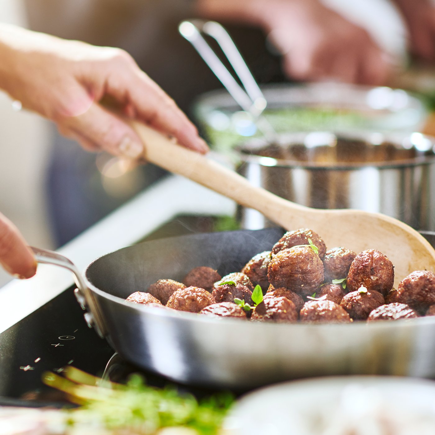 A frying pan full of meatless meatballs and chopped herbs being stirred by a person holding a wooden spoon.