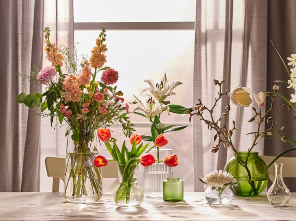 Devant la fenêtre habillée de rideaux fins d’une pièce ensoleillée, table avec différentes compositions florales présentées dans divers vases.
