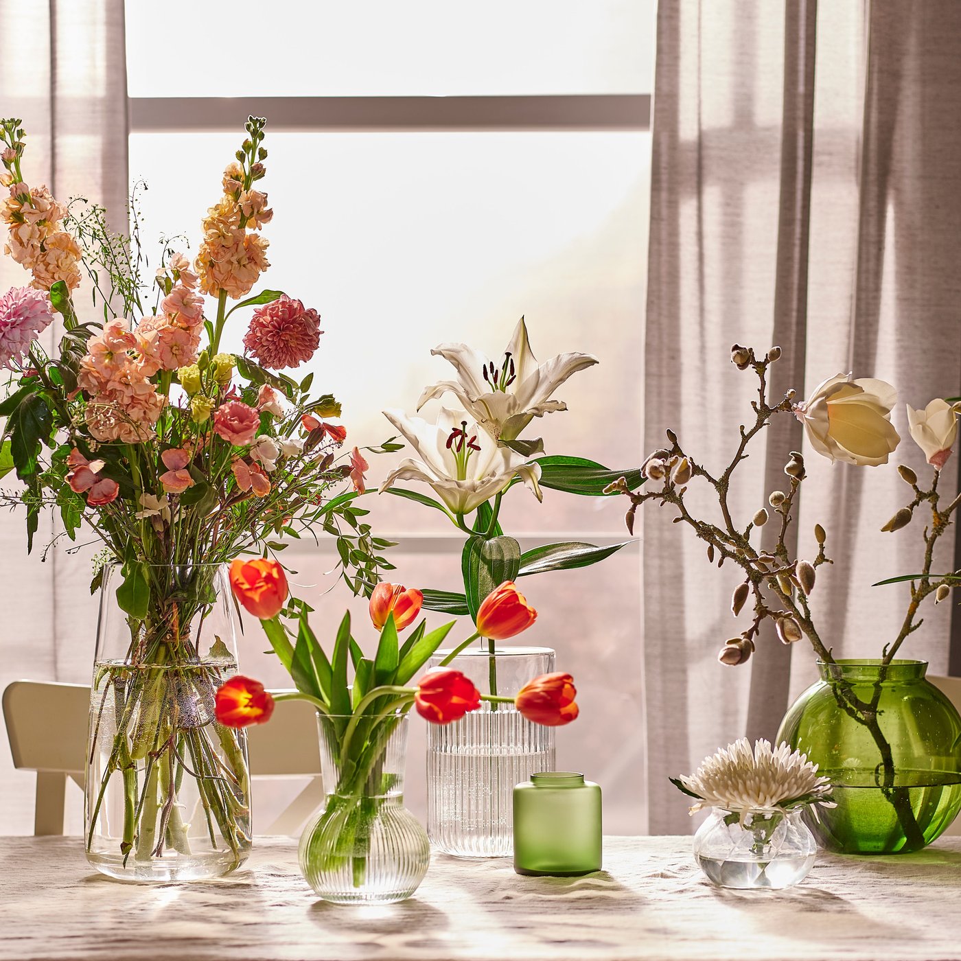 A table by a window with thin curtains with different flower arrangements in a variety of vases in a sunny room.