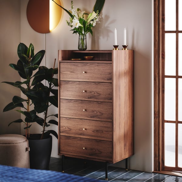 RÅDMANSÖ chest of 5 drawers in a warm walnut colour, standing between a green plant and the window in a bedroom.