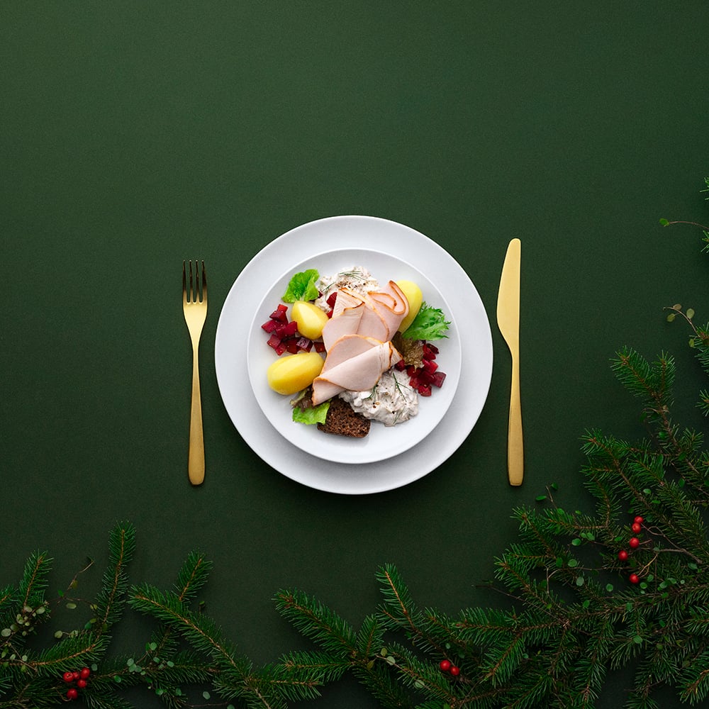 Vihreällä taustalla on näkyvissä kuusenoksia. Valkoiselle lautaselle on koottu Joulubuffetin tarjottavia, lautasen sivuilla on kultaiset aterimet, haarukka ja veitsi.