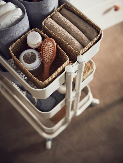 A white RÅSKOG trolley containing 4 storage boxes with baby towels, hairbrush, bottle and nappies on a brown carpet flooring.