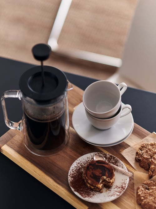 Prim-plan de bucătărie cu o placă de tocat maro din salcâm, care susține un aparat de preparat cafea și cești de cafea și farfurioare.