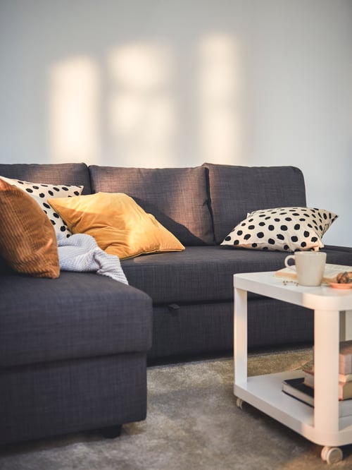 Wohnbereich mit einem FRIHETEN Eckbettsofa mit Aufbewahrung, zwei Kissen in ODDNY Kissenbezügen, einem TINGBY Beistelltisch mit Rollen und einem STOENSE Teppich.