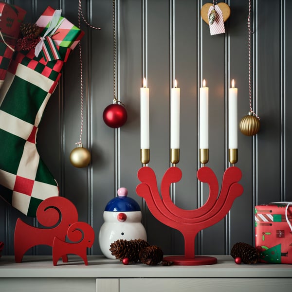 En röd ljusstake med fyra tända vita ljus på en hylla, omgiven av hängande ornament och juldekorationer.