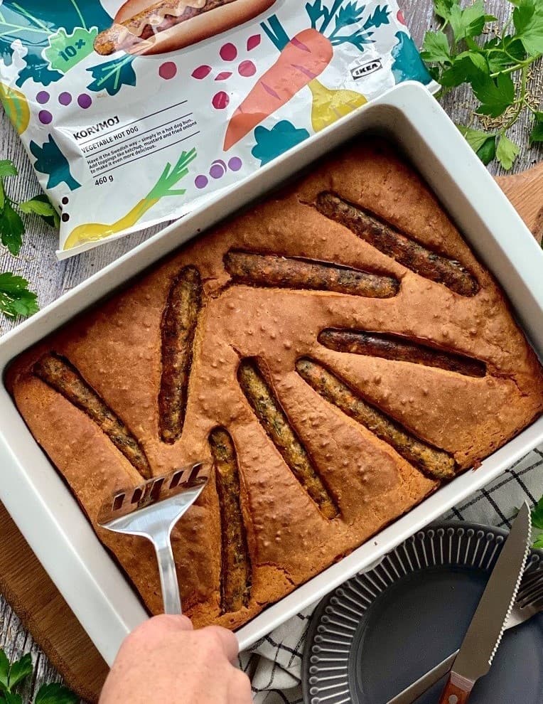 Une personne sert un plat cuit au four garni de morceaux ressemblant à des saucisses, entouré de légumes verts frais, avec un emballage de produit IKEA en arrière-plan.