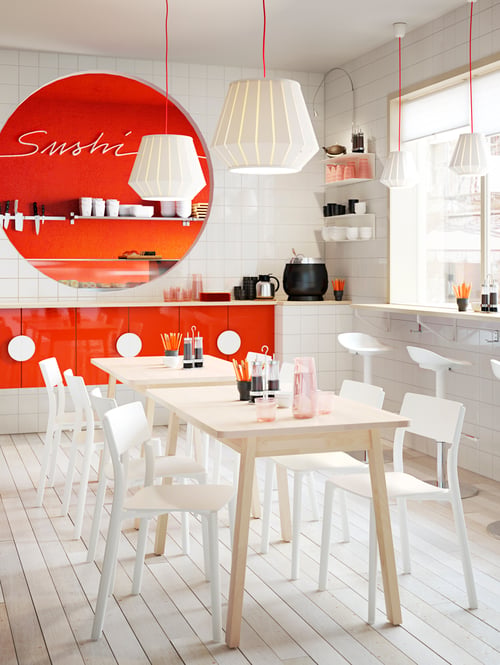 Dining area with a rectangular table, white chairs, white bar stools, white lamp shades, and orange wall cabinets.