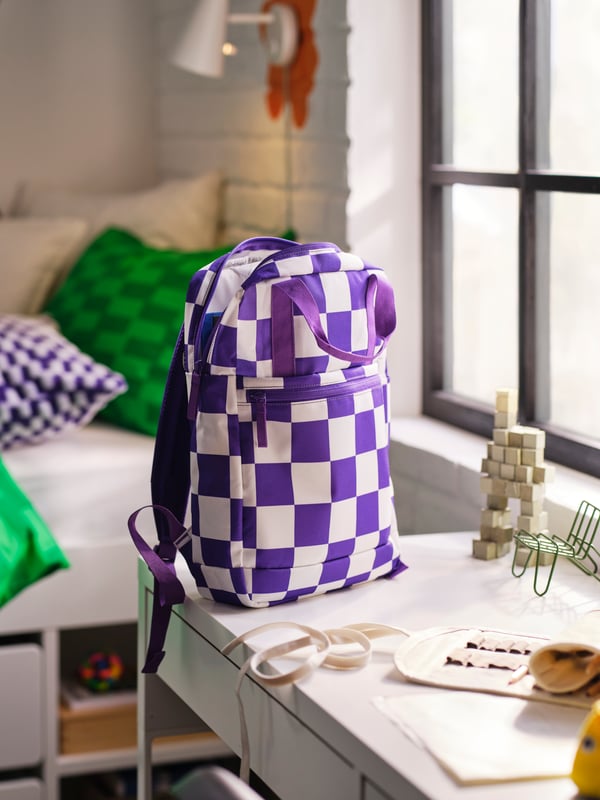 Es ist ein Raum voller unterschiedlicher Accessoires zu sehen. In der Mitte befindet sich eine Tasche, die ein kariertes Muster in den Farben Lila und Weiss hat.