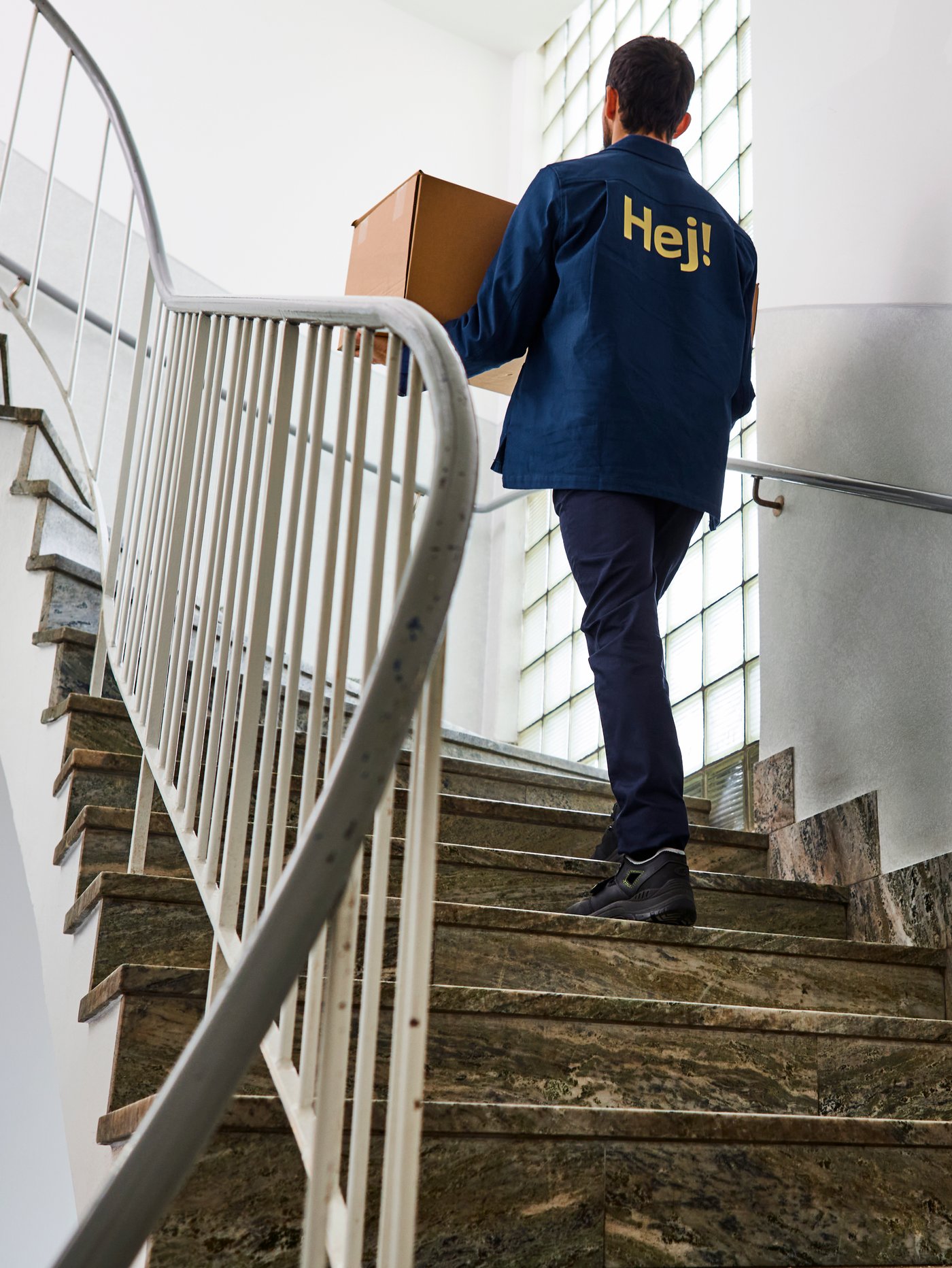 A man in a blue IKEA jacket with the word "Hej!" carries a carton box up a flight of stairs.