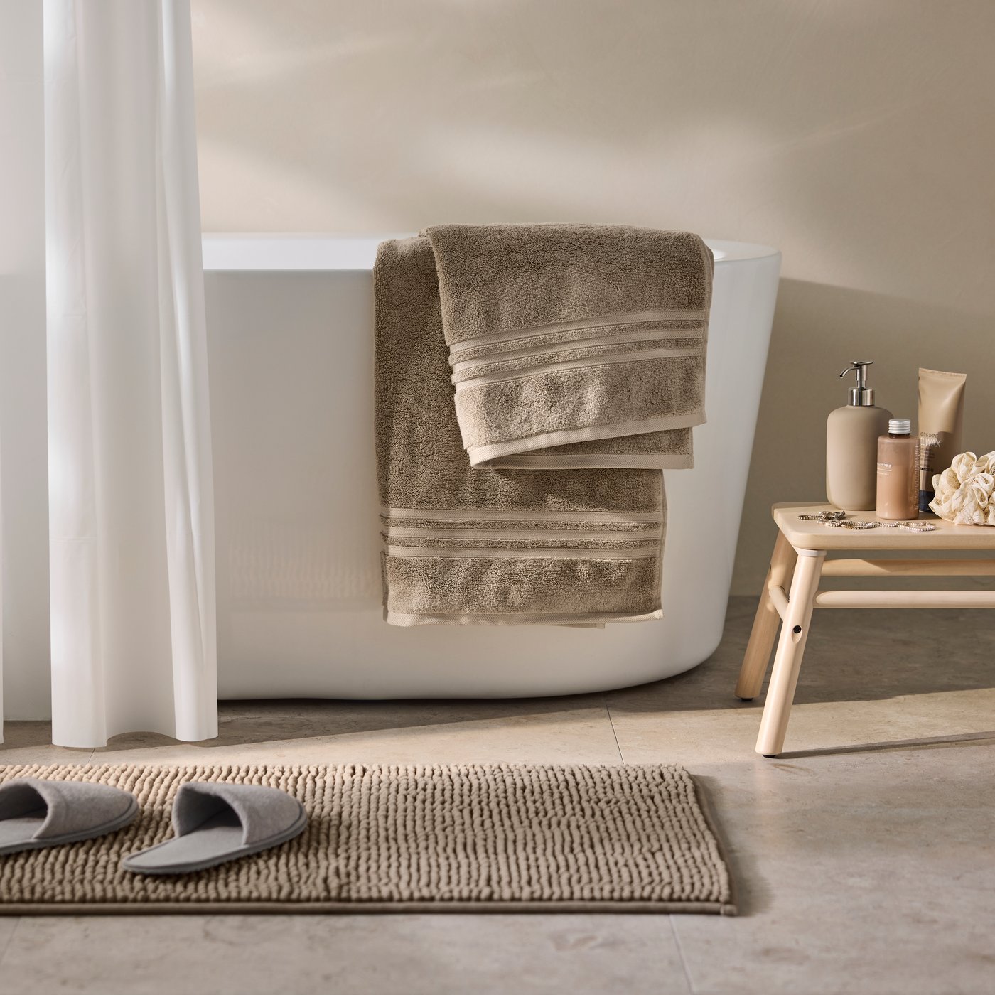 A bathroom featuring a beige BROKGLIM bath towel hanging on a white bath tub and a wooden stool nearby.