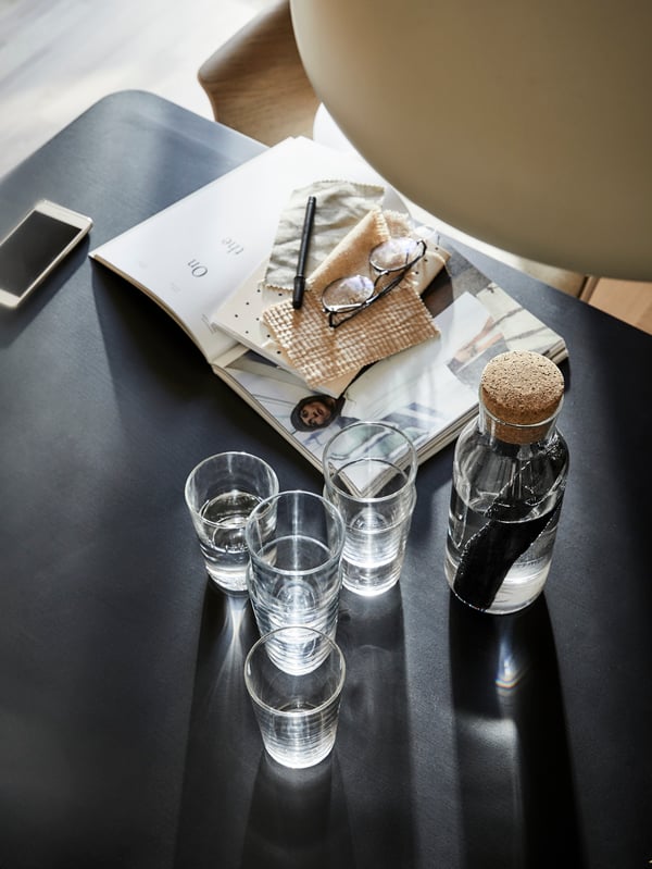 Drikkeglas og en glaskaraffel med korkprop står på et sort bord, ved siden af et åbent magasin og en telefon.