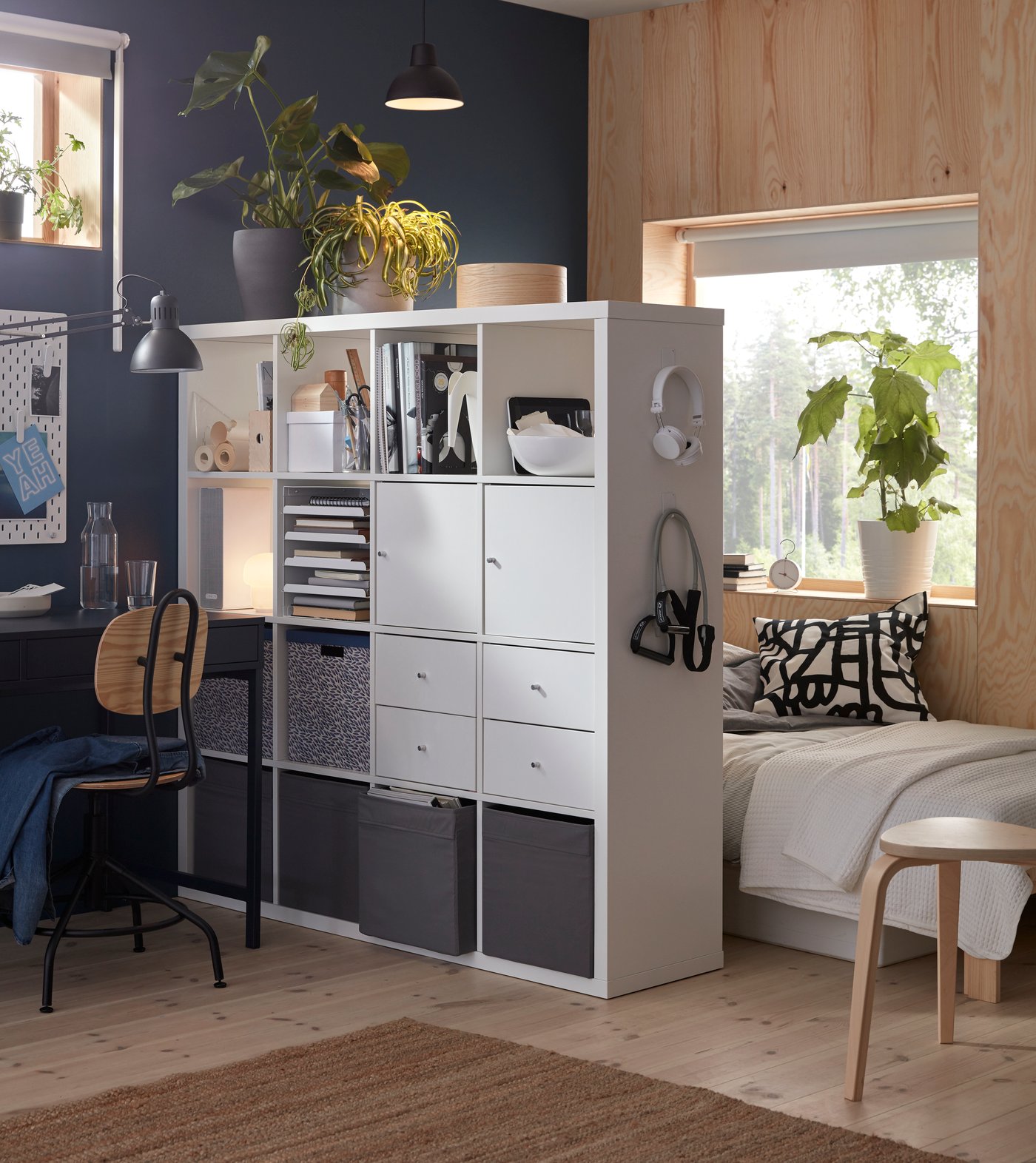 A room divided with a white KALLAX shelving unit with four inserts and DRÖNA boxes, creating a sleep zone and a workspace.