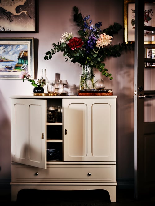 Un salon dans lequel on retrouve une armoire beige, un vase noir, des vases en verre, des fleurs artificielles, des cadres et des objets décoratifs.