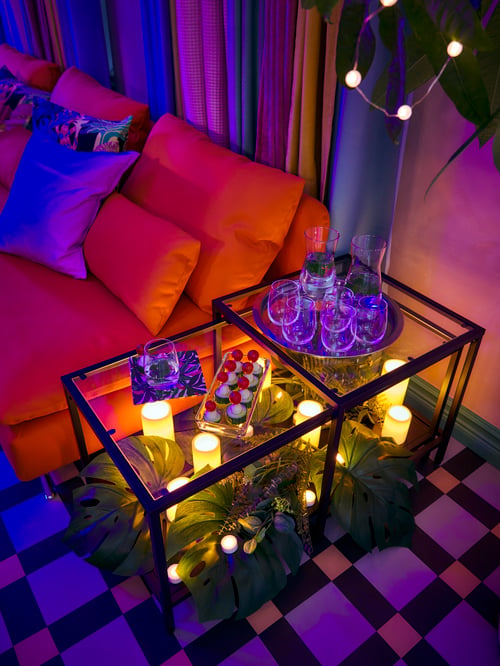 Un salon avec un canapé et deux tables gigognes en acier avec un plateau en verre trempé, des bougies à DEL et un plateau avec des verres.