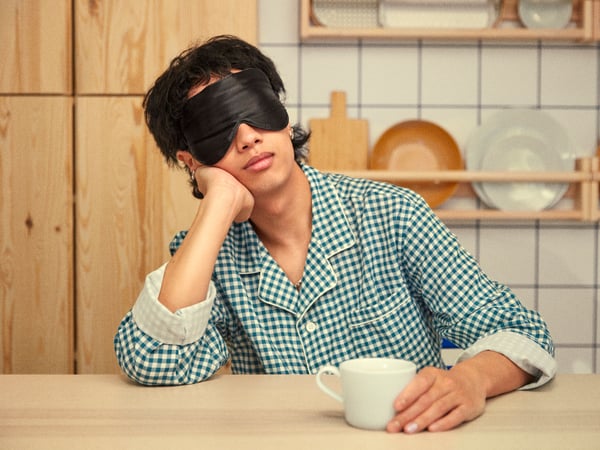 A young man dressed in a pyjama is sitting at the kitchen table with an eye mask on and a mug in his hand.