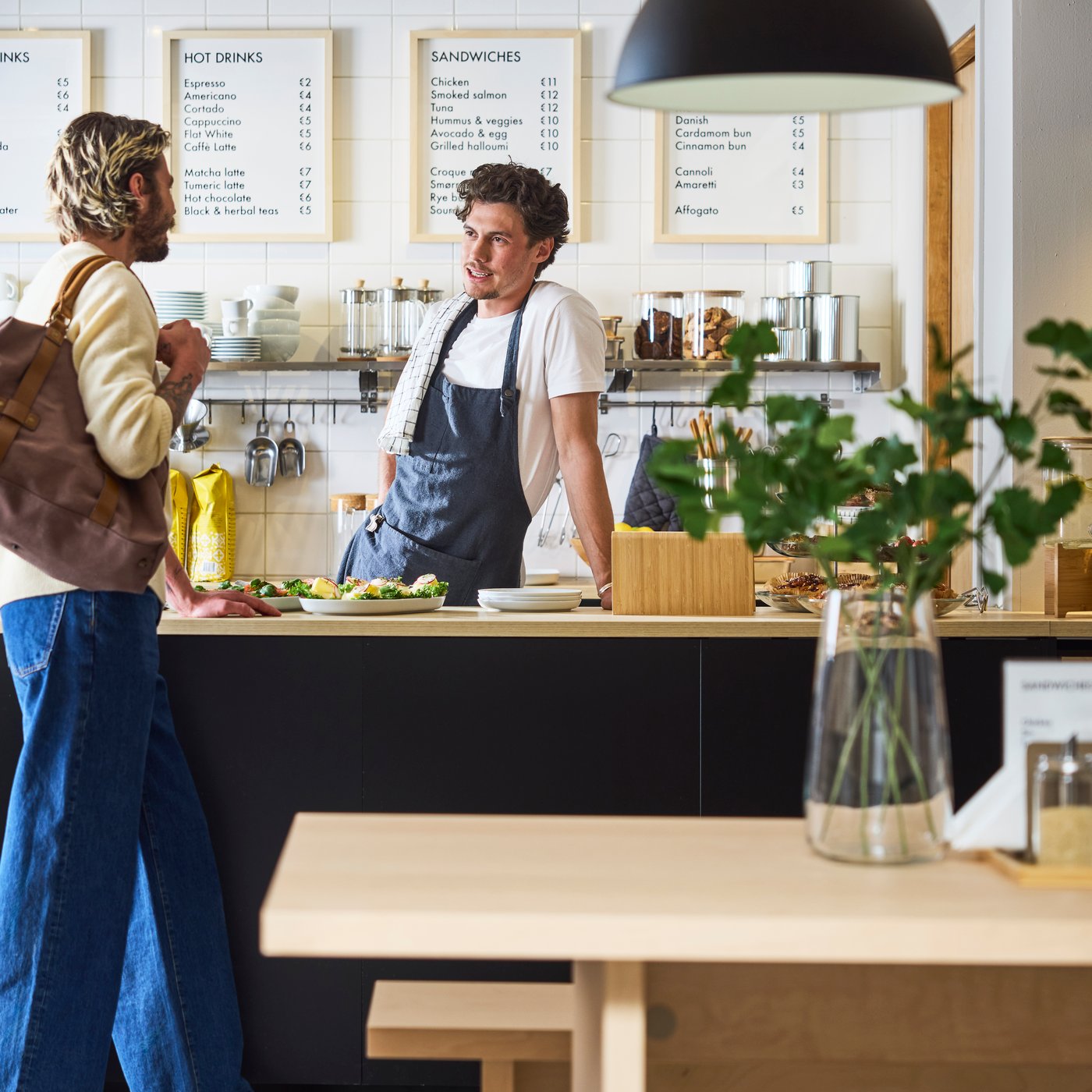 Barista bakom cafédisken pratar med kunden; menytavlor, koppar och bakverk syns i den ljusa, moderna interiören.